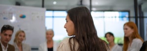 Woman with brown hair sitting at a table with a group of coworkers leading a learning exercise.