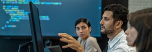 Three coworkers looking at a monitor with a larger screen in the background displaying code. 