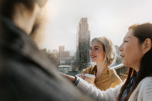 Woman clutching a coffee talking with friends with a city skyline in the background