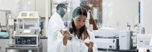Two scientists in white lab coats working in a research laboratory, with one using a pipette over a sample tray in the foreground.