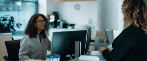 A smiling woman in glasses sitting at an office desk, interacting with a colleague