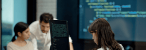 Three colleagues collaborating in a tech workspace, with code displayed on computer monitors and a large screen in the background.