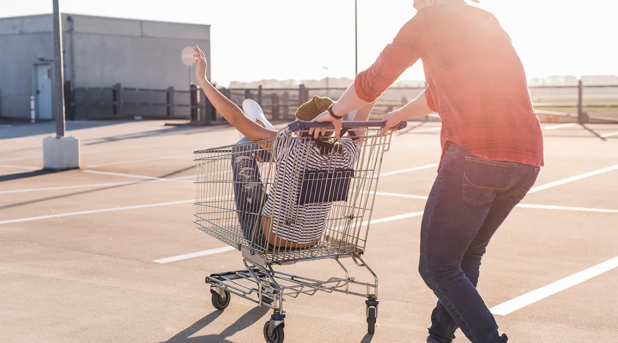 A man pushes a woman in a shopping cart through an empty parking lot.